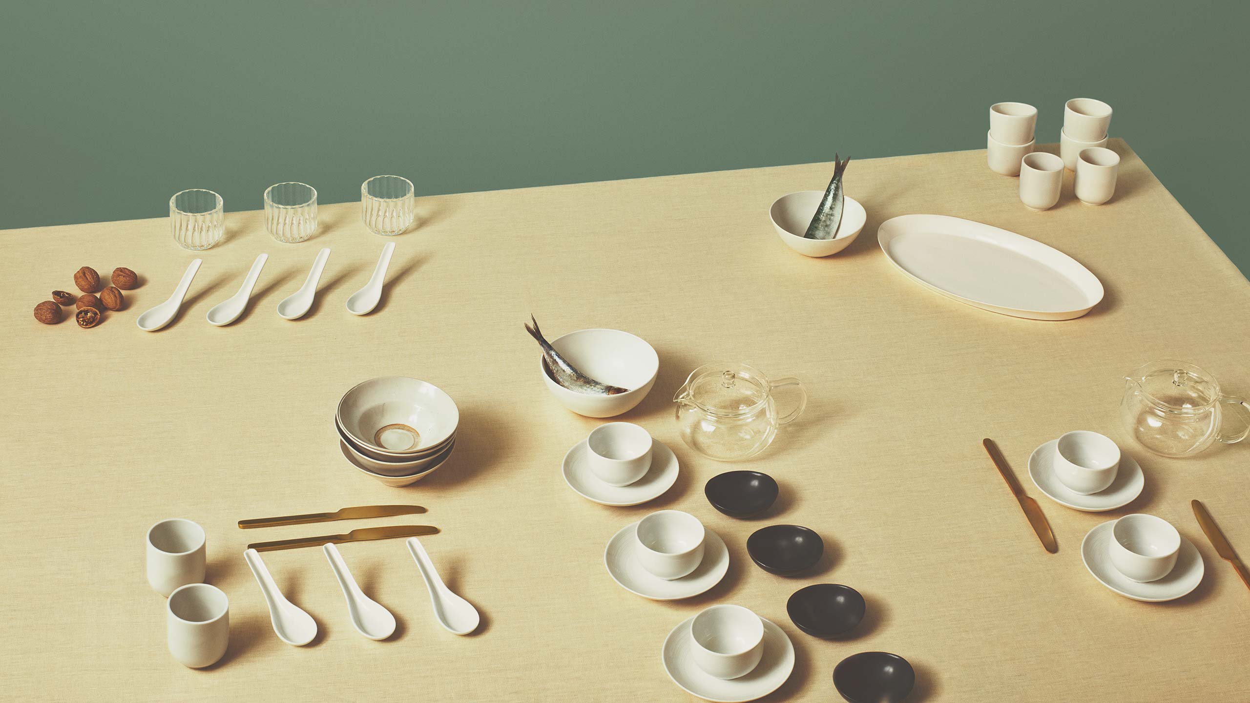 A table set for a meal, with cups, teapots, soup bowls and cutlery on an oatmeal-coloured tablecloth.