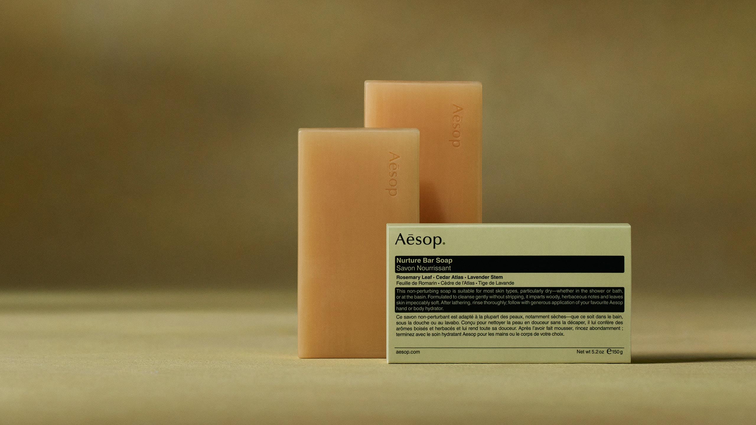 Aesop Nurture bar soaps placed horizontally behind the packaging in dark green textured background