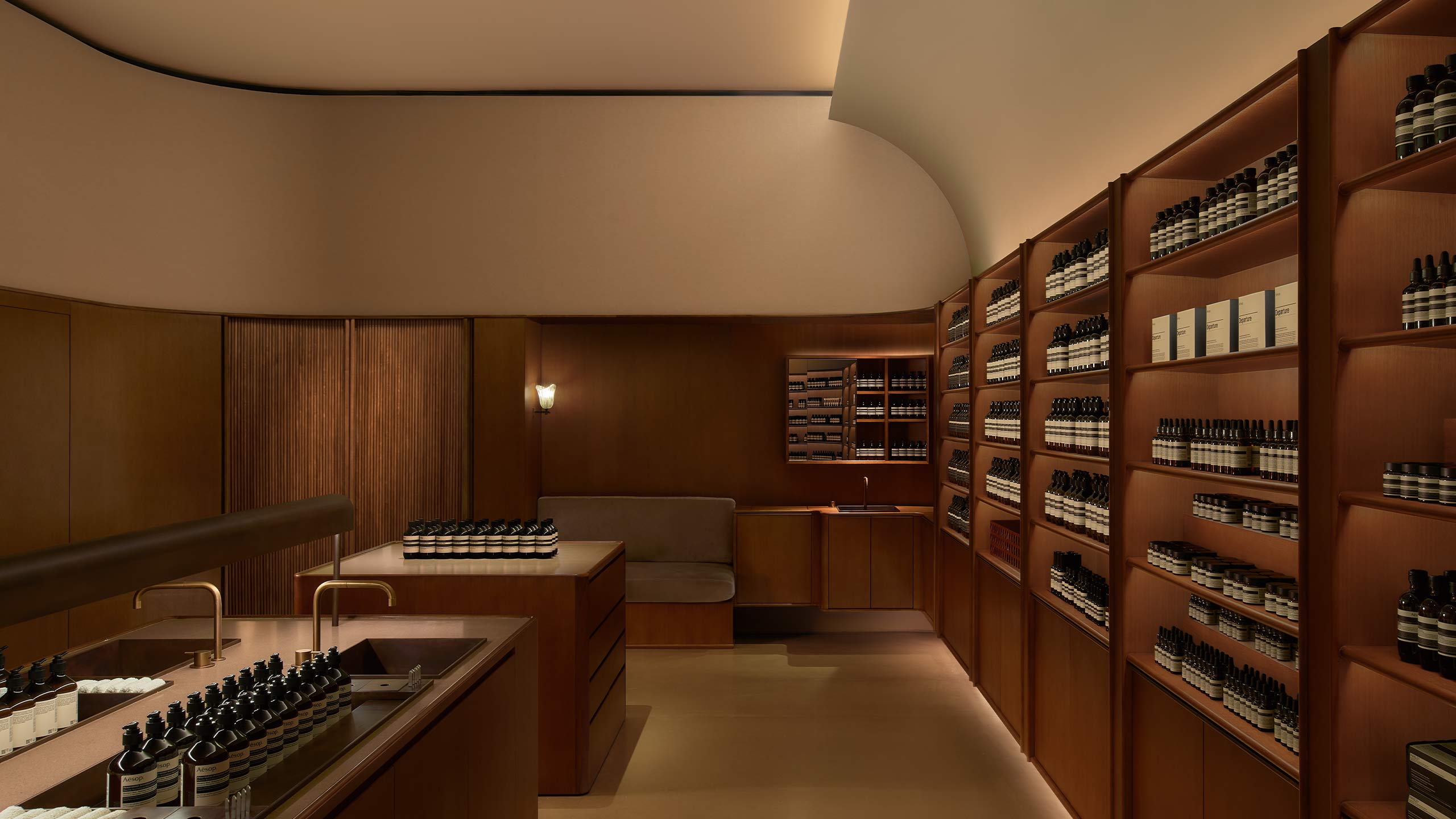A quartet of bronze basins occupies the centre, with Aesop full range products sheathed on walnut timber cabinetry, and a velvet sofa at the edge.