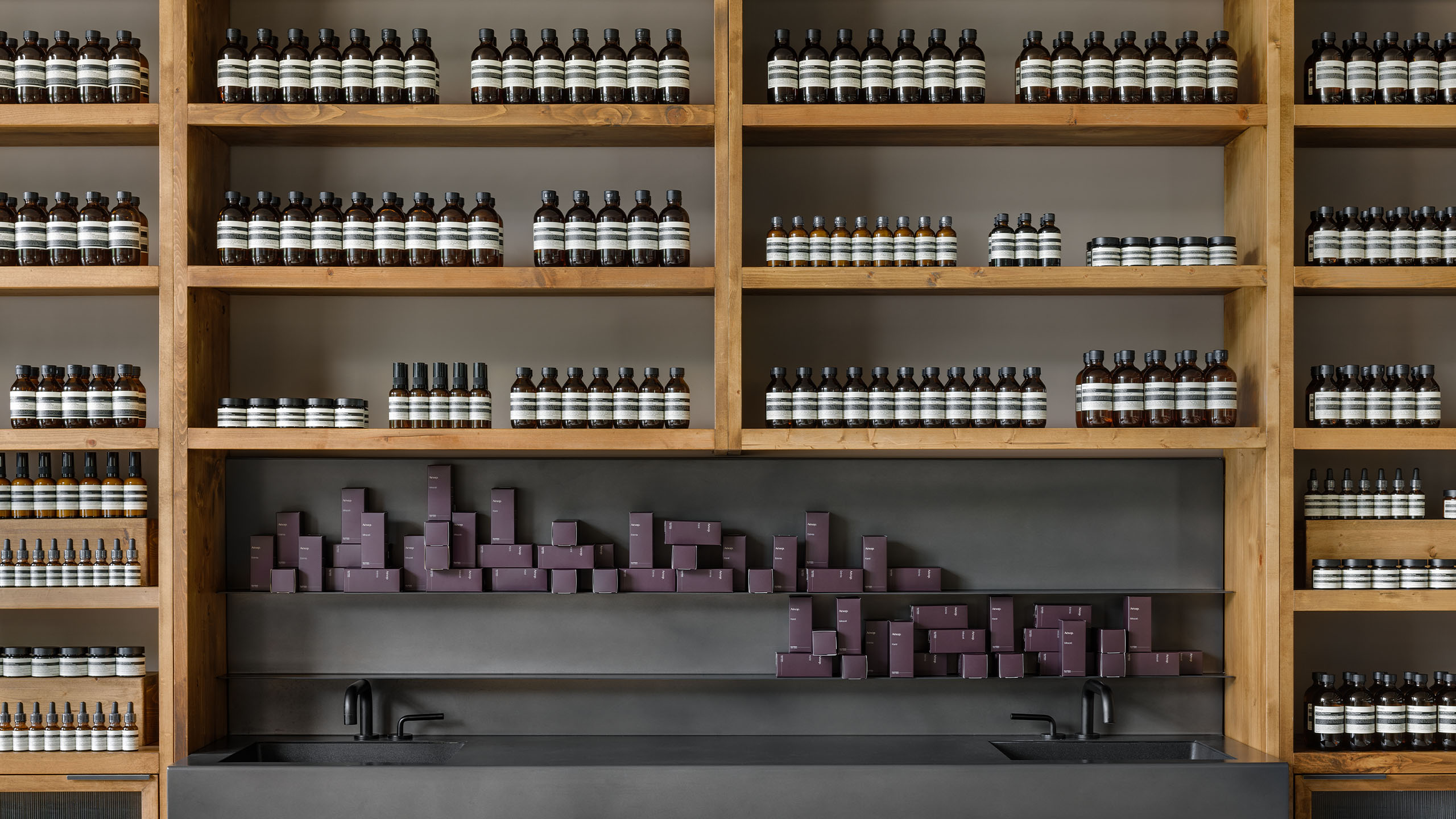 A dark grey sink accompanied by wooden shelves holding Aesop fragrance collection.