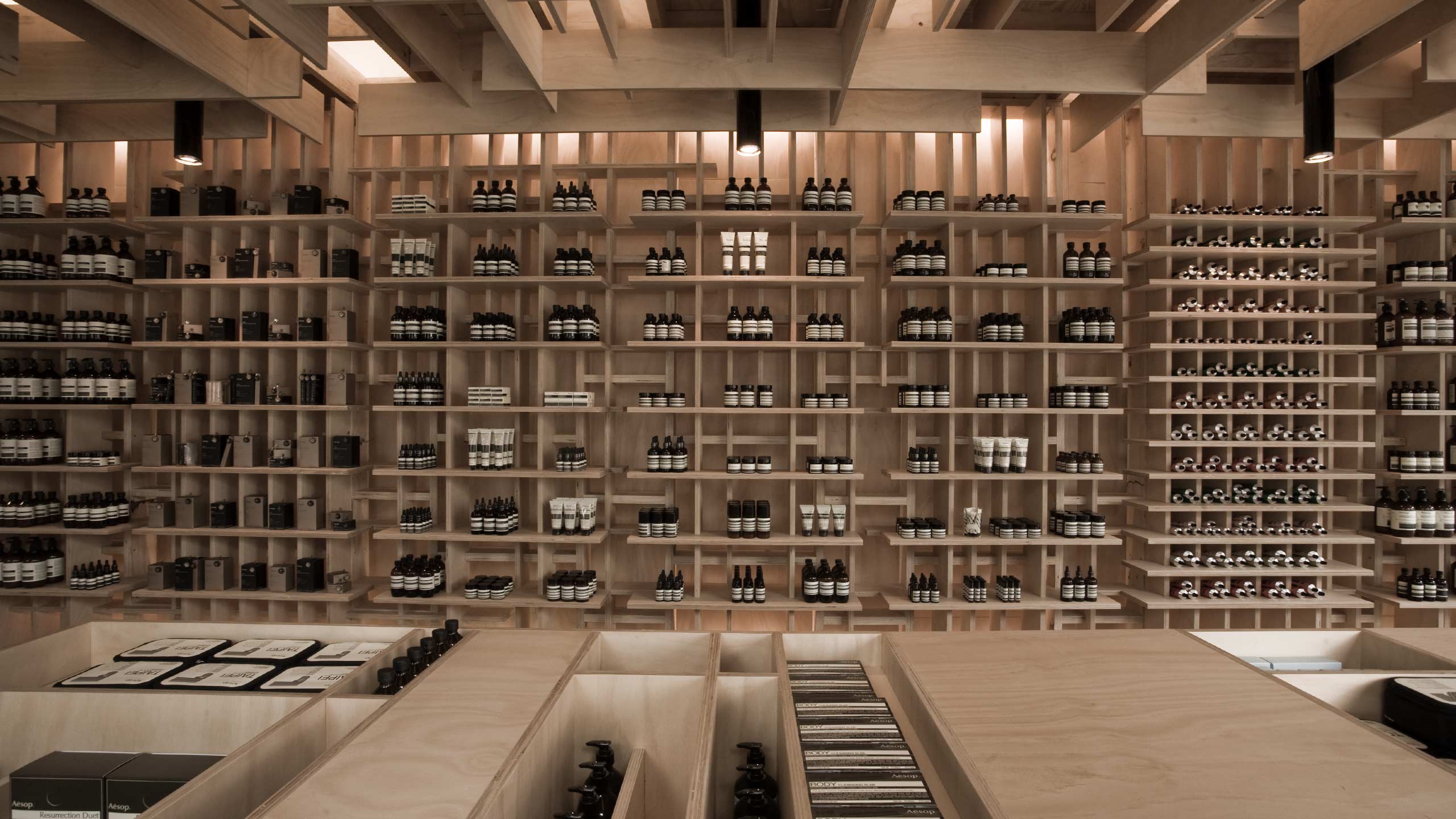 Black Aesop bottles line an intricate framework of birch shelving. A wooden counter-top is in the foreground.