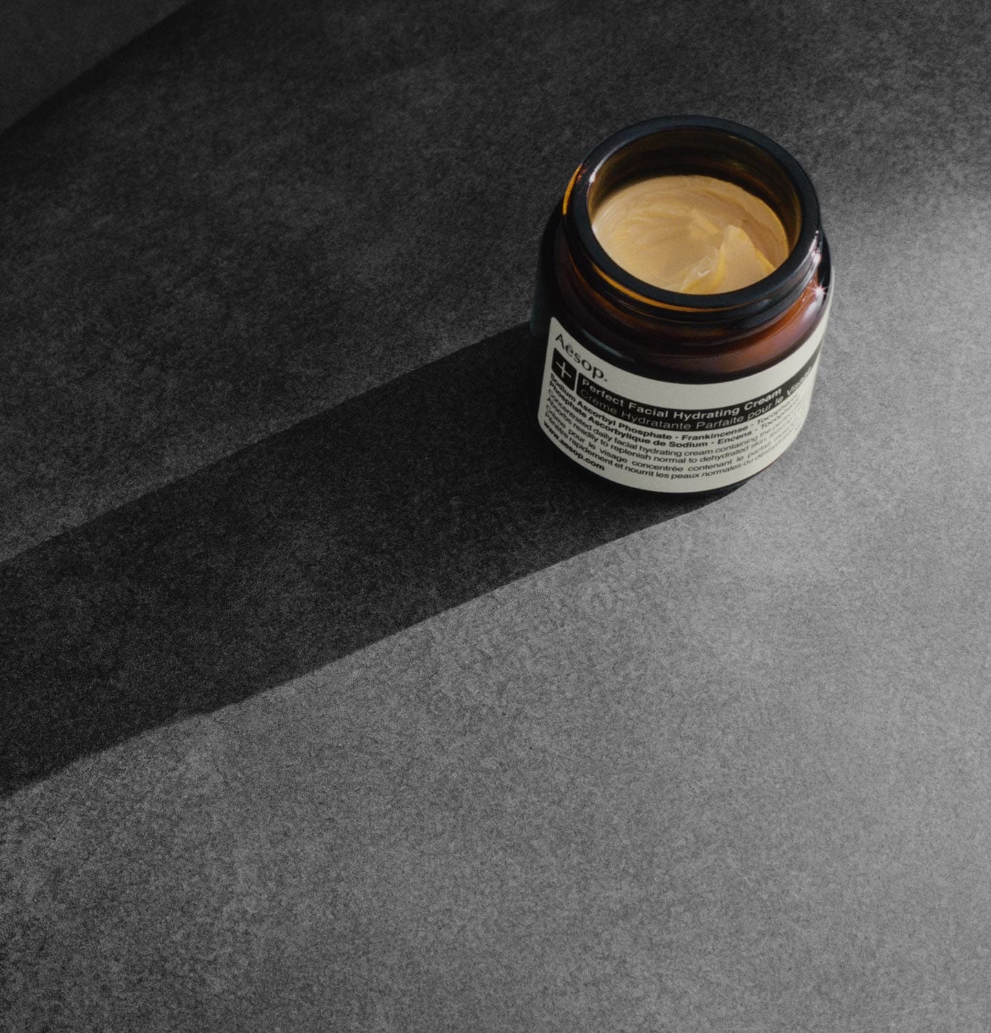 A jar of Perfect Facial Hydrating Cream sits open on a concrete surface to reveal a smooth, buttery cream. 