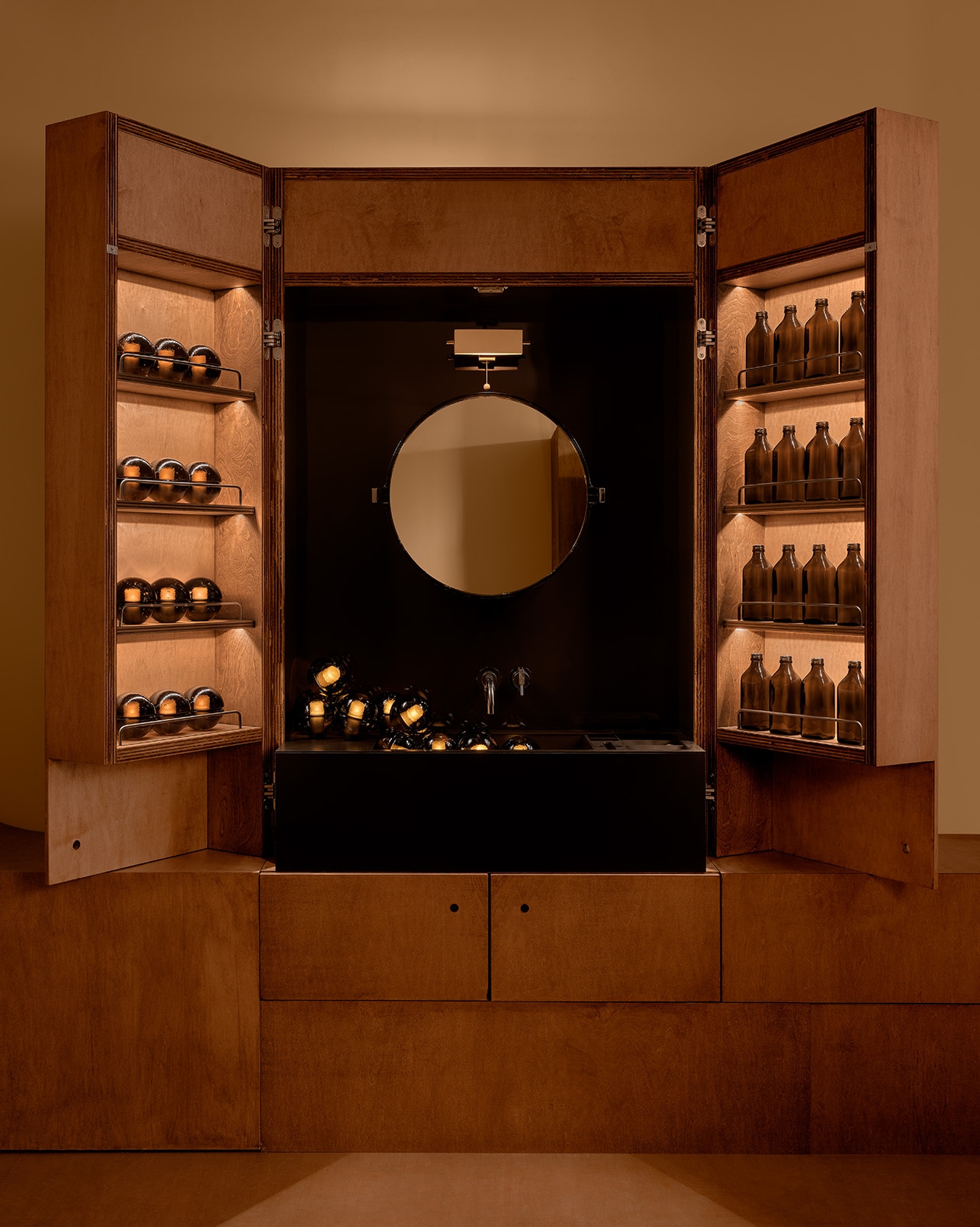 warmly lit armoire with round mirror in center of black panel, with illuminated wood doors open featuring amber glass bottles and illuminated amber glass orb lamps on shelves