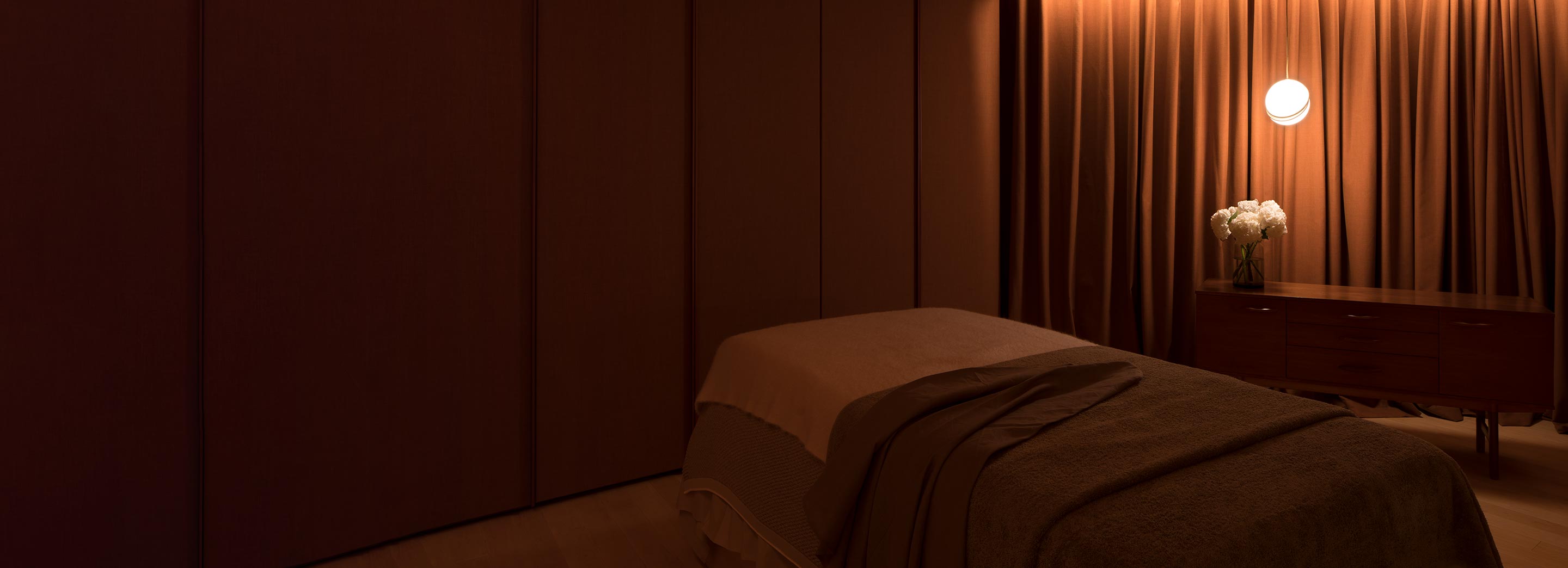 A dim room with a massage bed and a wooden cabinet with a bouquet of flowers placed on top. A orange coloured lamp hanging on the cabinet.