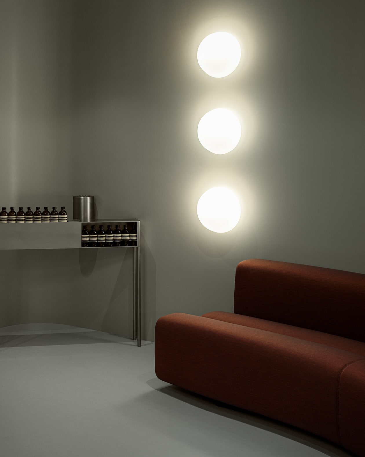 photo of grey room with aesop product on steel table, three circle shaped lights mounted vertically on wall, and red couch