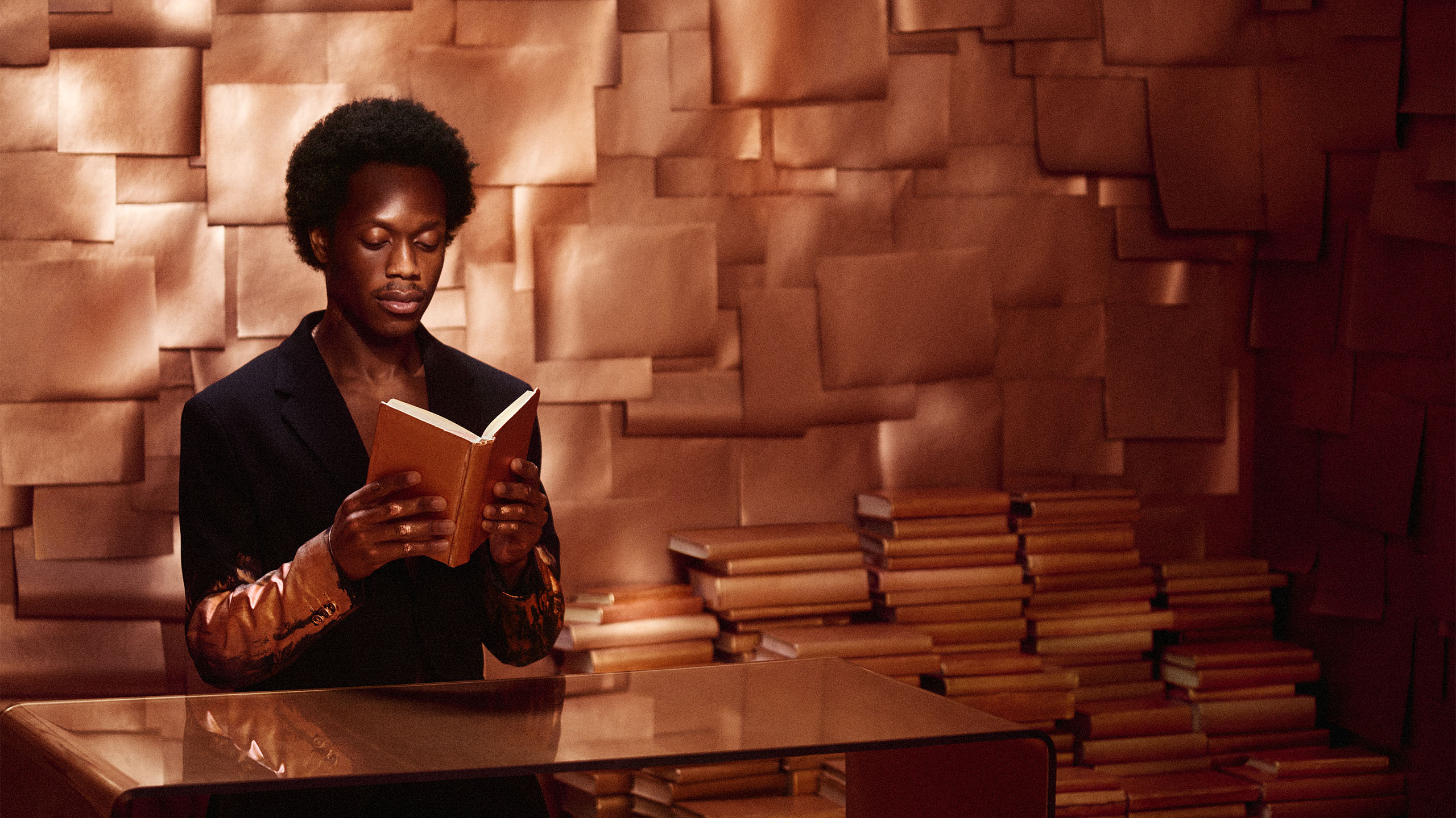 person reading book sitting at a table in bronze lighting in front of stacks of books and wall papered with sheets of paper of different sizes