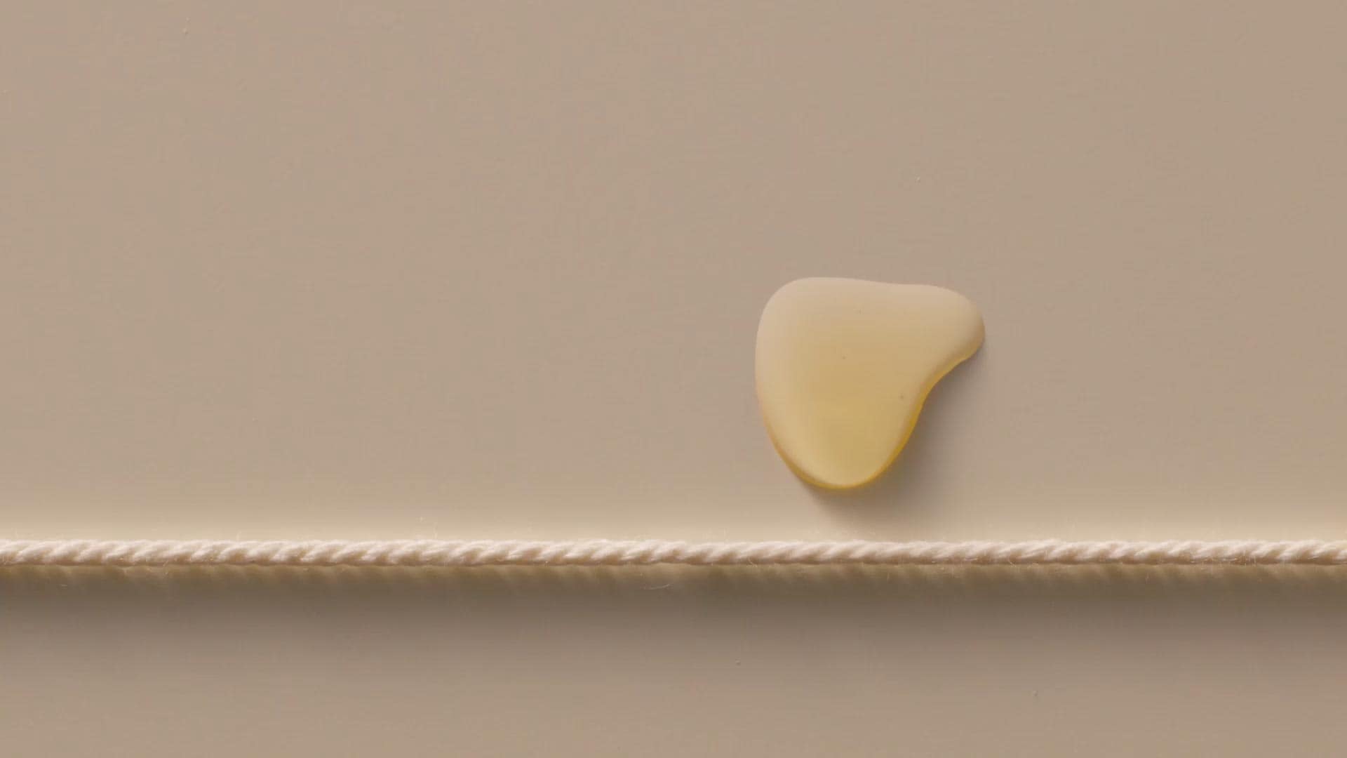 A drop of serum dripping on a beige surface above a white thread