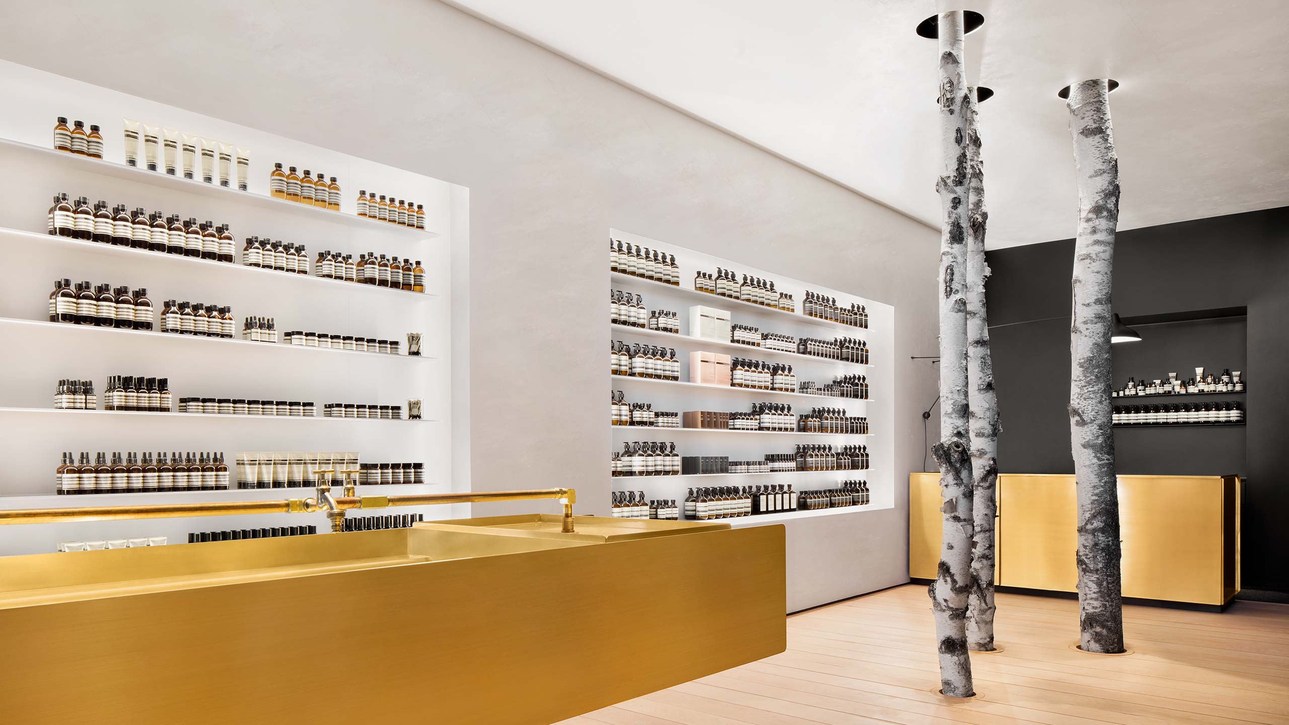Three tall tree trunks acting as supporting beams within a modernist an Aesop store interior; fitted with brass counters and sinks against ice-black walls.