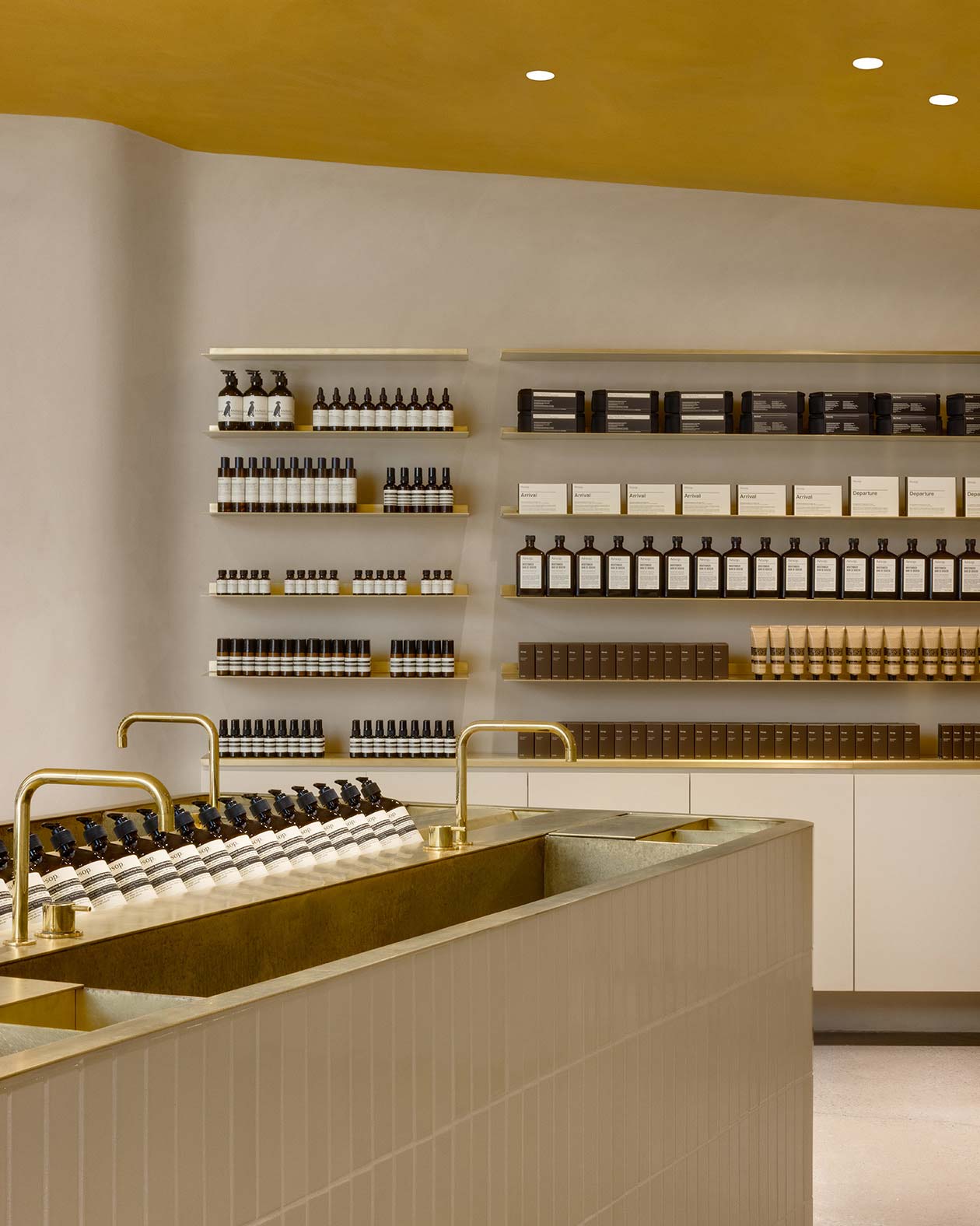 A sink fitted with a brass tap and countertop in the foreground; shelves of Aesop product are neatly arranged on the shelves in the background. 