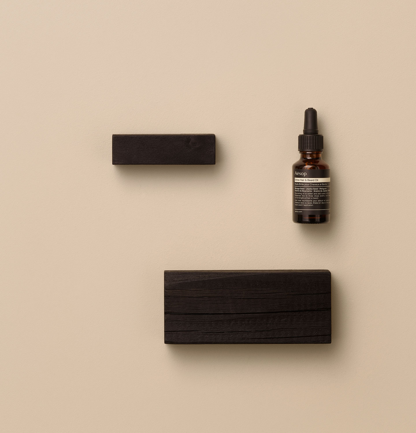 Shine Hair & Beard Oil de Aesop colocado junto a un objeto de madera oscura en una superficie beige.