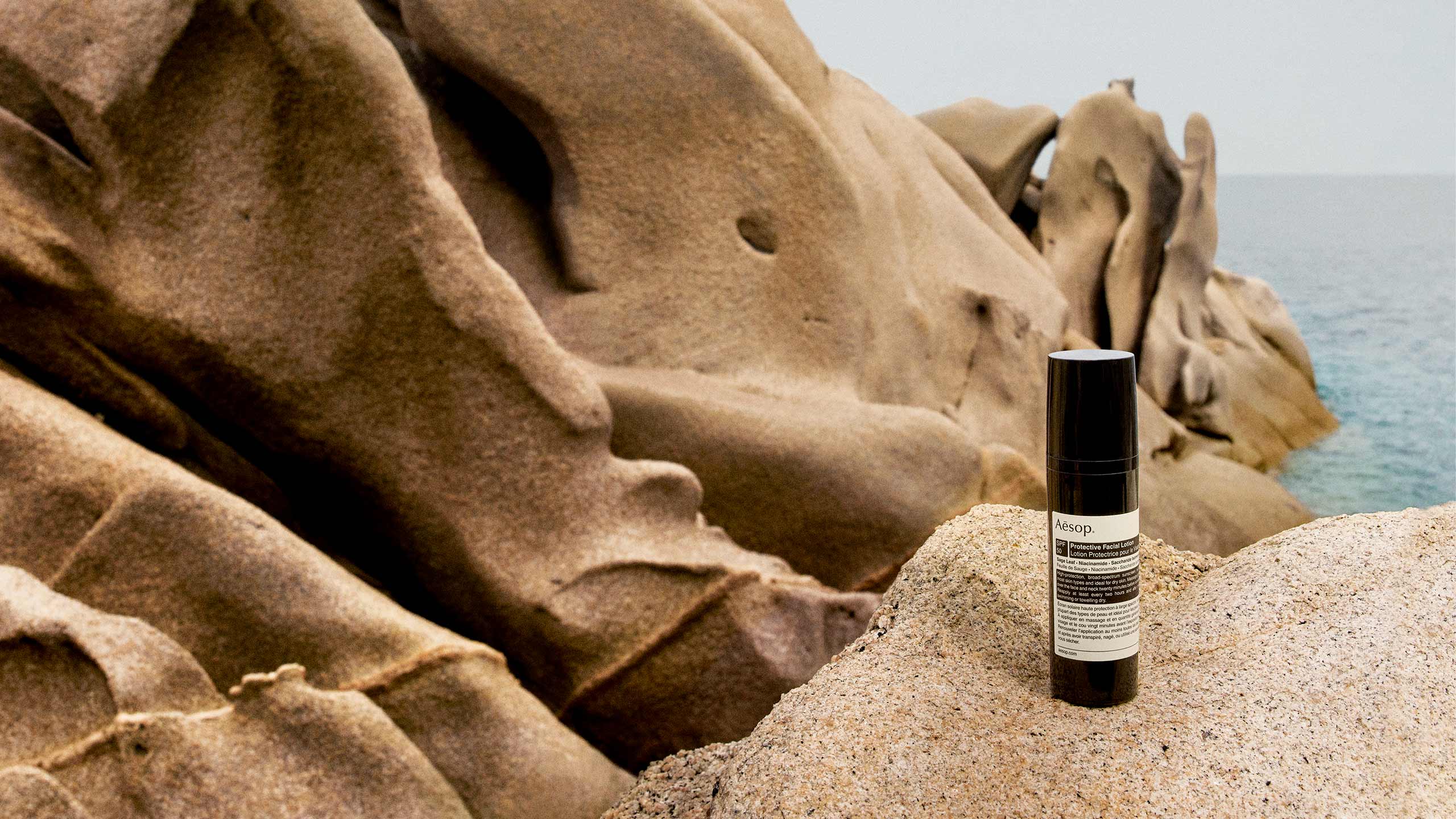 A bottle of Protective Facial Lotion SPF50 resting on a rocky shoreline.