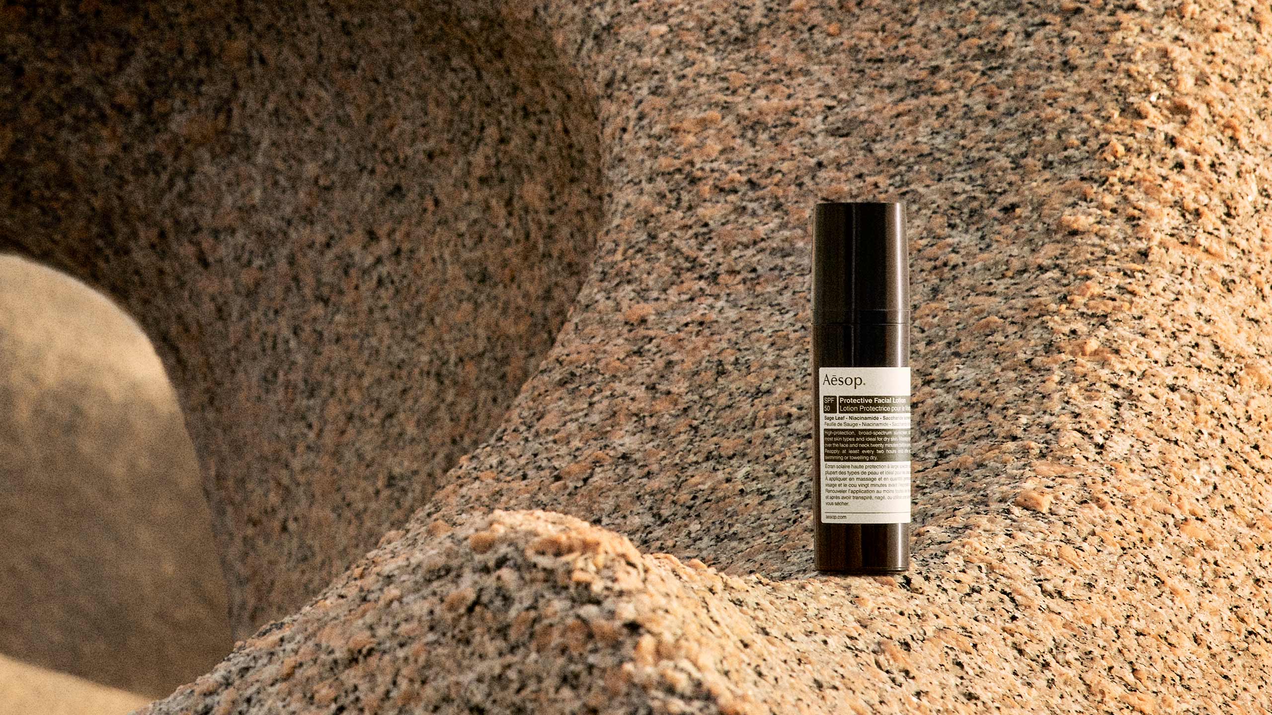 A bottle of Protective Facial Lotion SPF50 resting on a rocky shoreline.