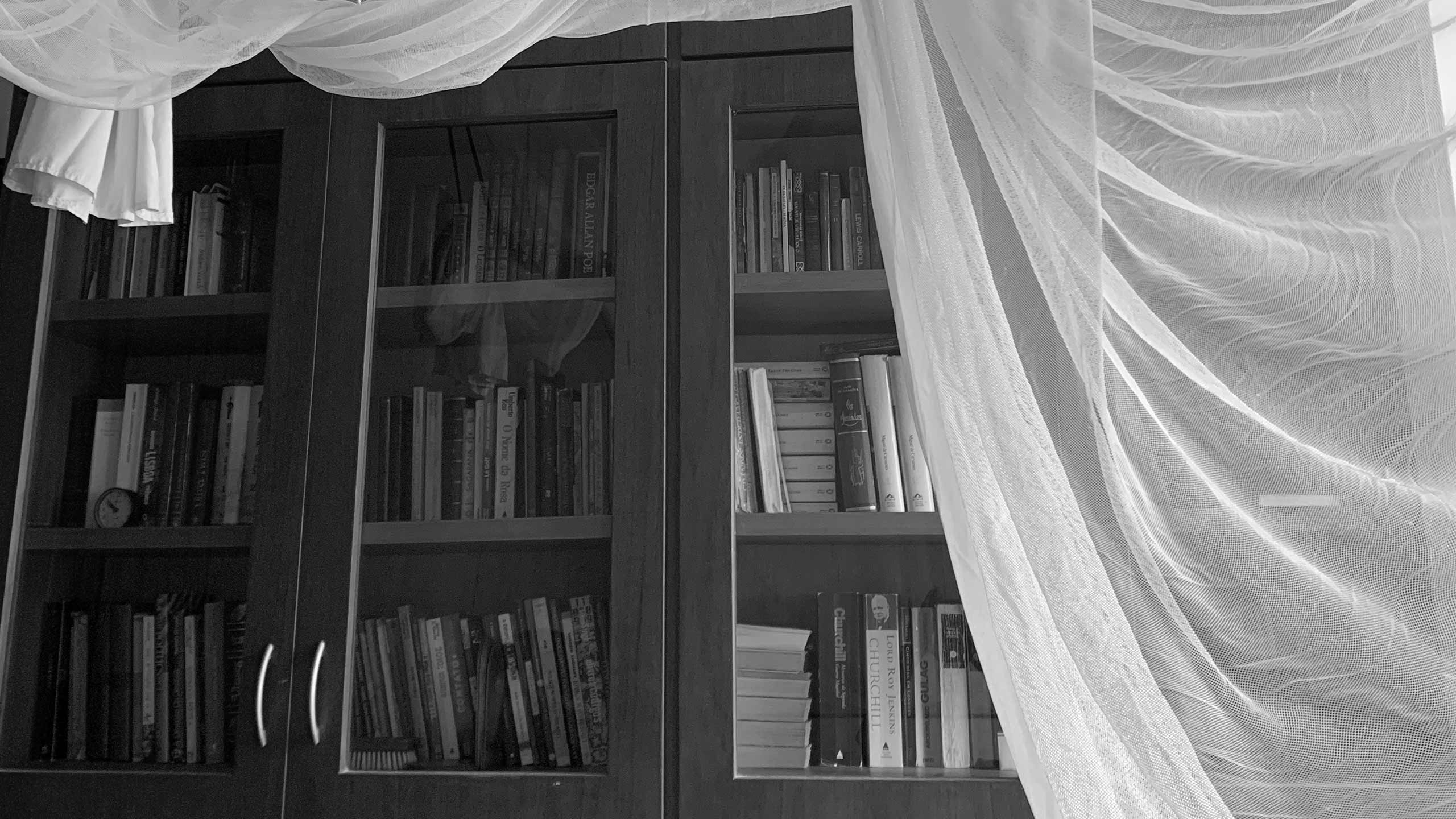 An antique wooden bookshelf next to curtains blowing in the wind.