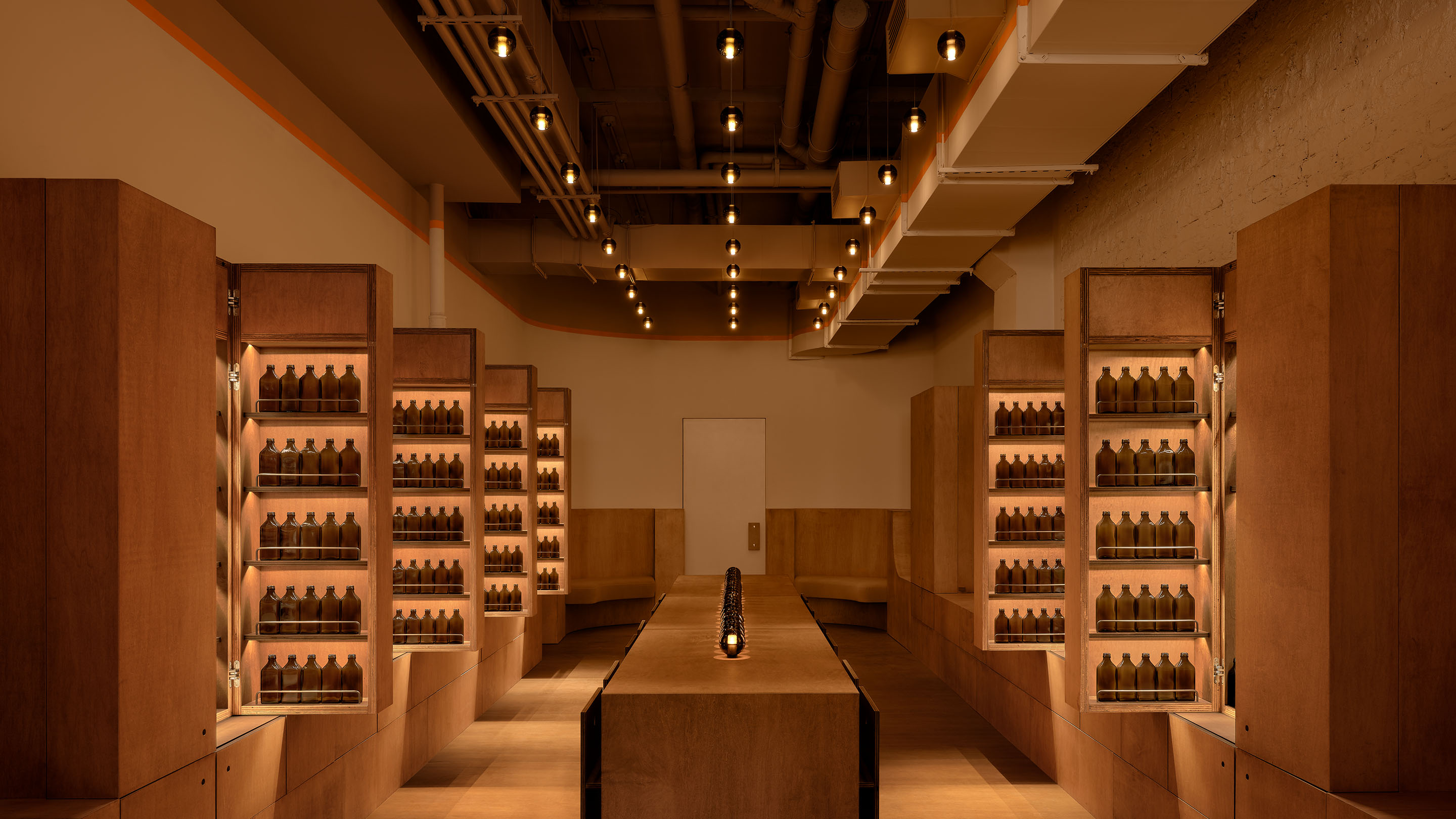interior store shot of warmly lit retail space with center counter unit flanked by walkways and product on shelves, paneled in wood and lit overhead and on the counter by amber glass orb lamps