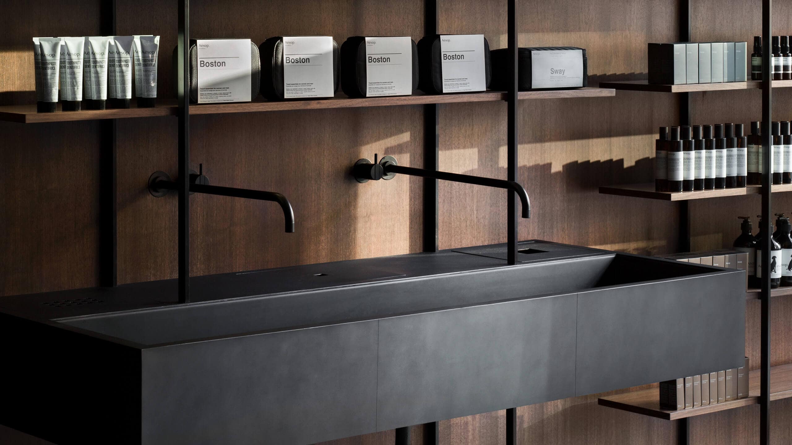 A close-up of black steel taps and basins with Aesop Travel Kits arranged on the shelf above.