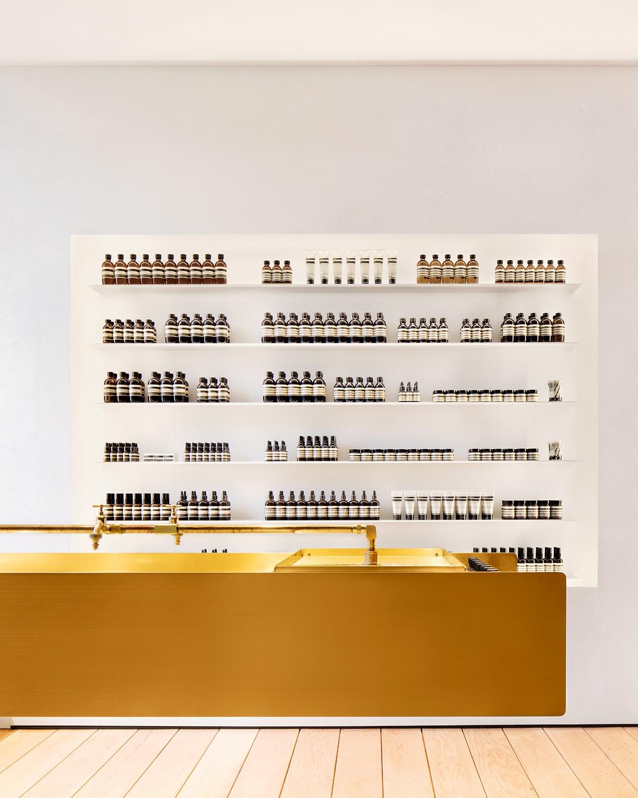Pure white walls stocked with Aesop product in an orderly fashion; a brass rectangular sink is positioned in-front.