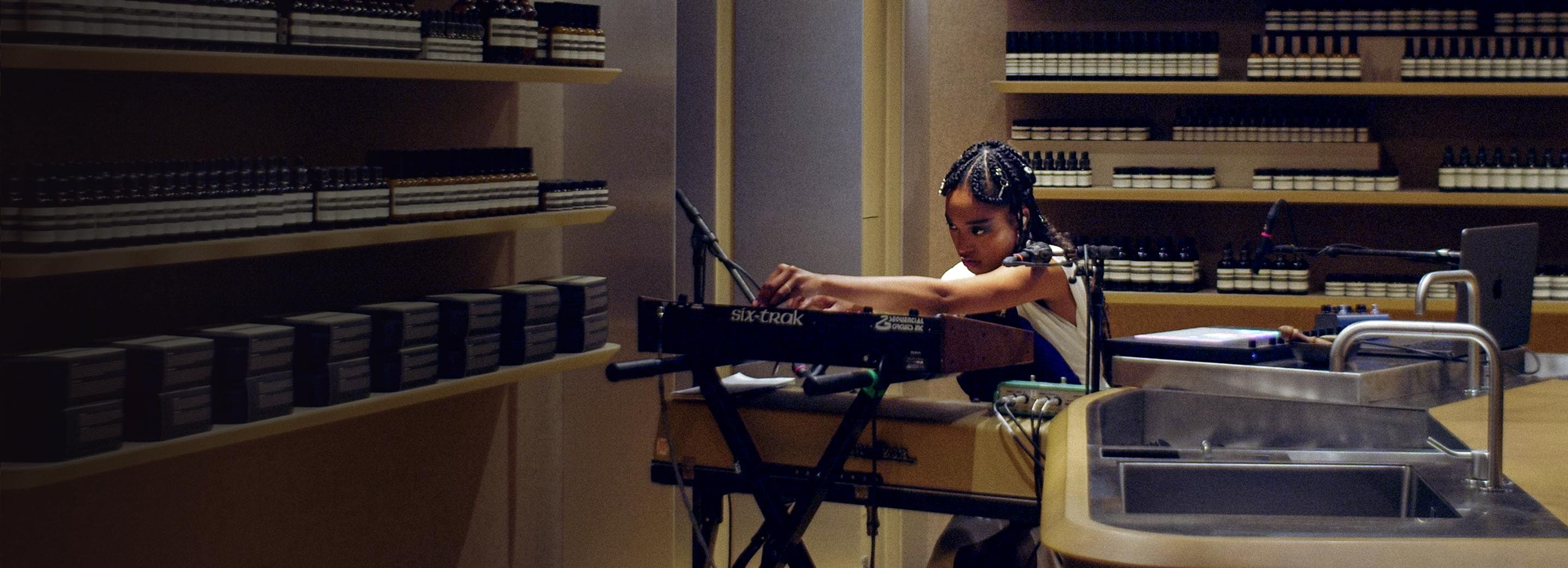 Kiala Ogawa operating a synthesizer surrounded by amber glass bottles in an Aesop store.