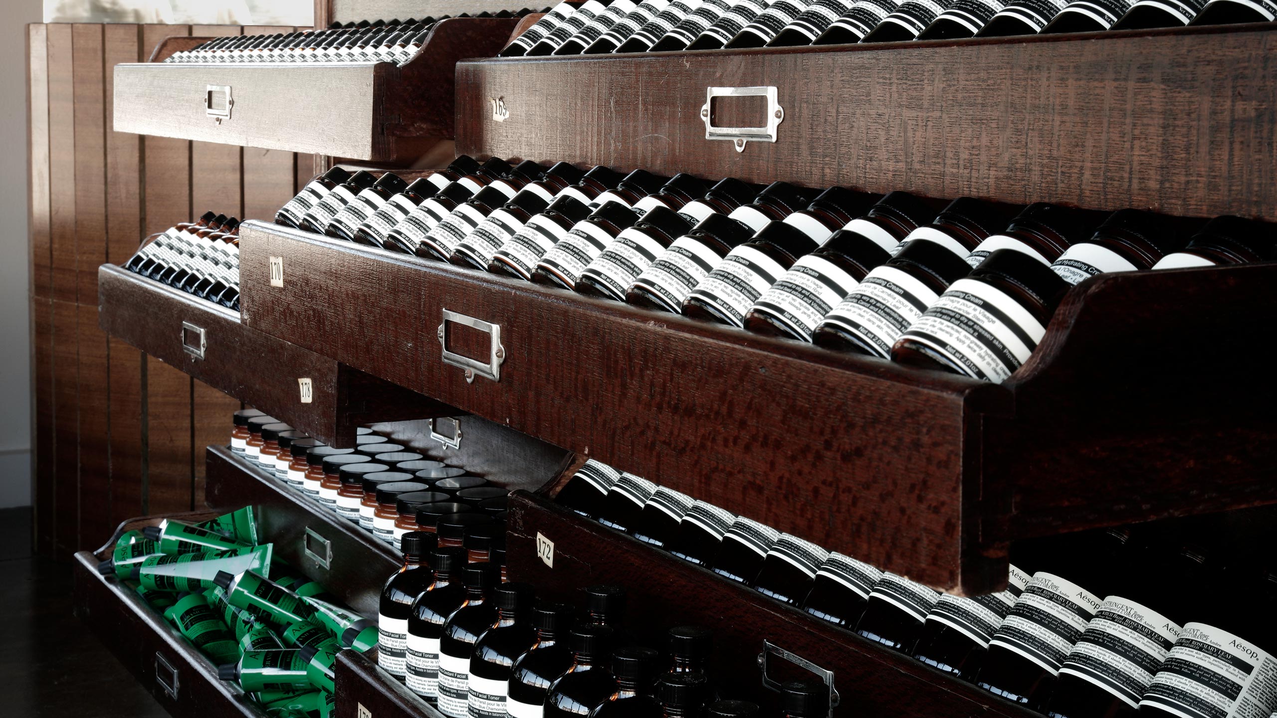 Dark oak archival drawers are filled with different Aesop products in dark bottles.