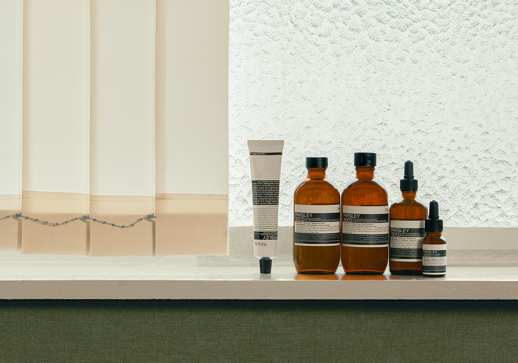 Aesop range of Parsely Seed products in a row, lined on a shelf next to a set of window blinds.