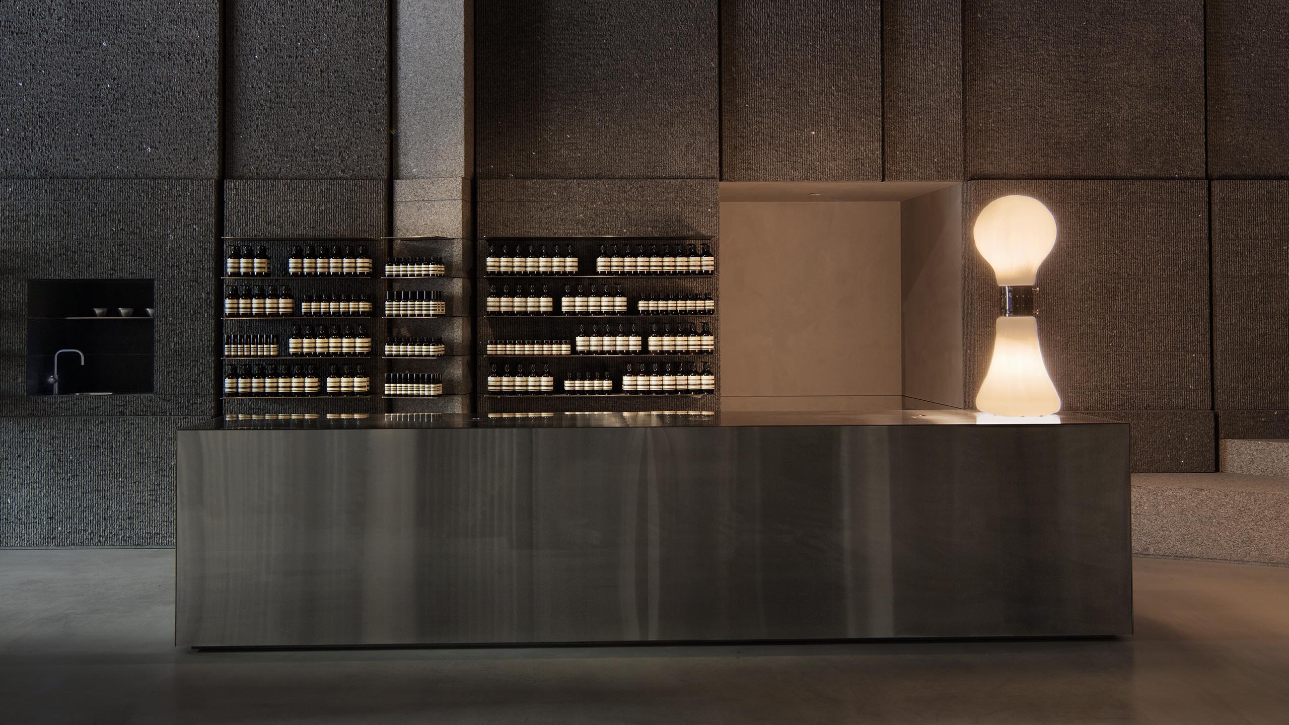 A steel basin with in-front of a concrete wall displaying amber bottles of Aesop Product