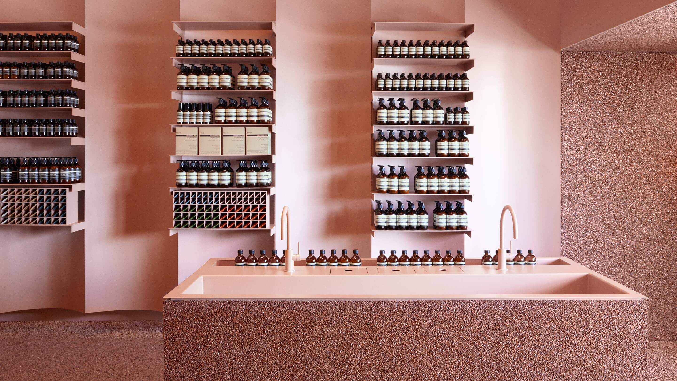 Salmon pink shelves holding rows of Aesop product; speckled pink sinks sit in-front. 