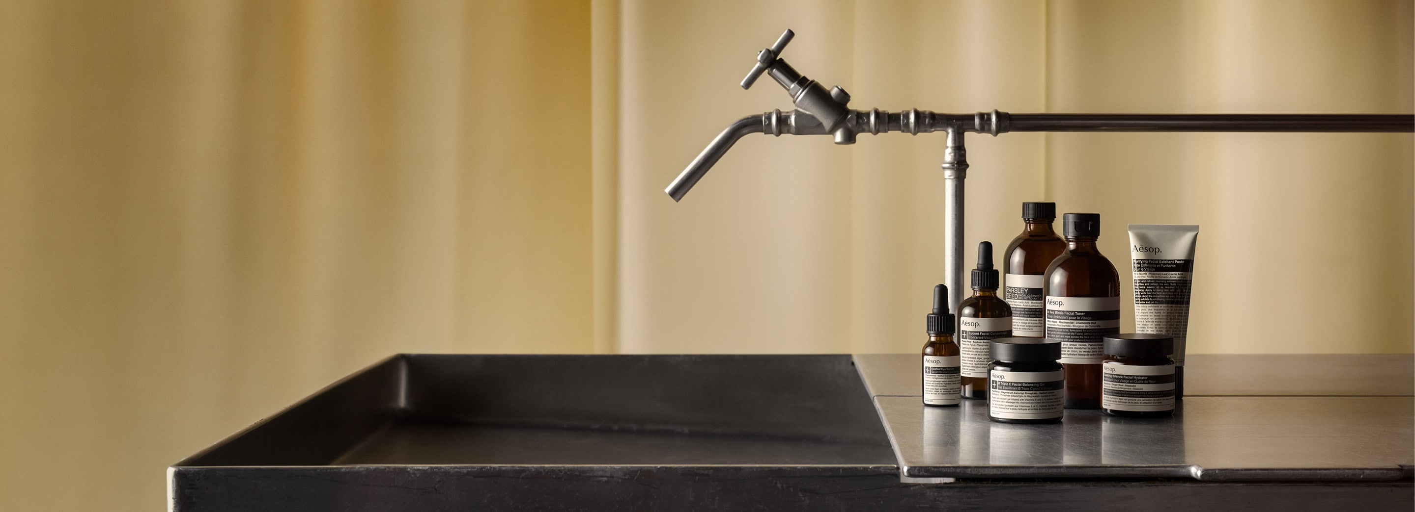 Skin Care products resting on a metallic surface, beside a sink and faucet, in front of a yellow curtain.
