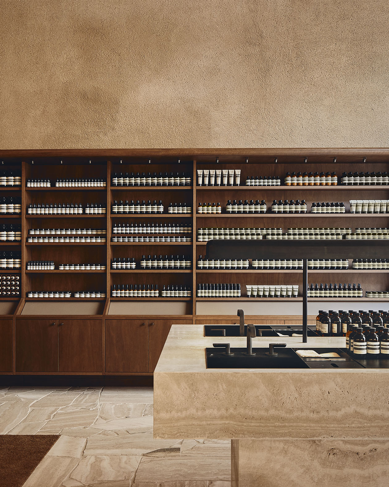 Travertine sink sits in the foreground; dark walnut wooden shelves holding Aesop product featured in the background.