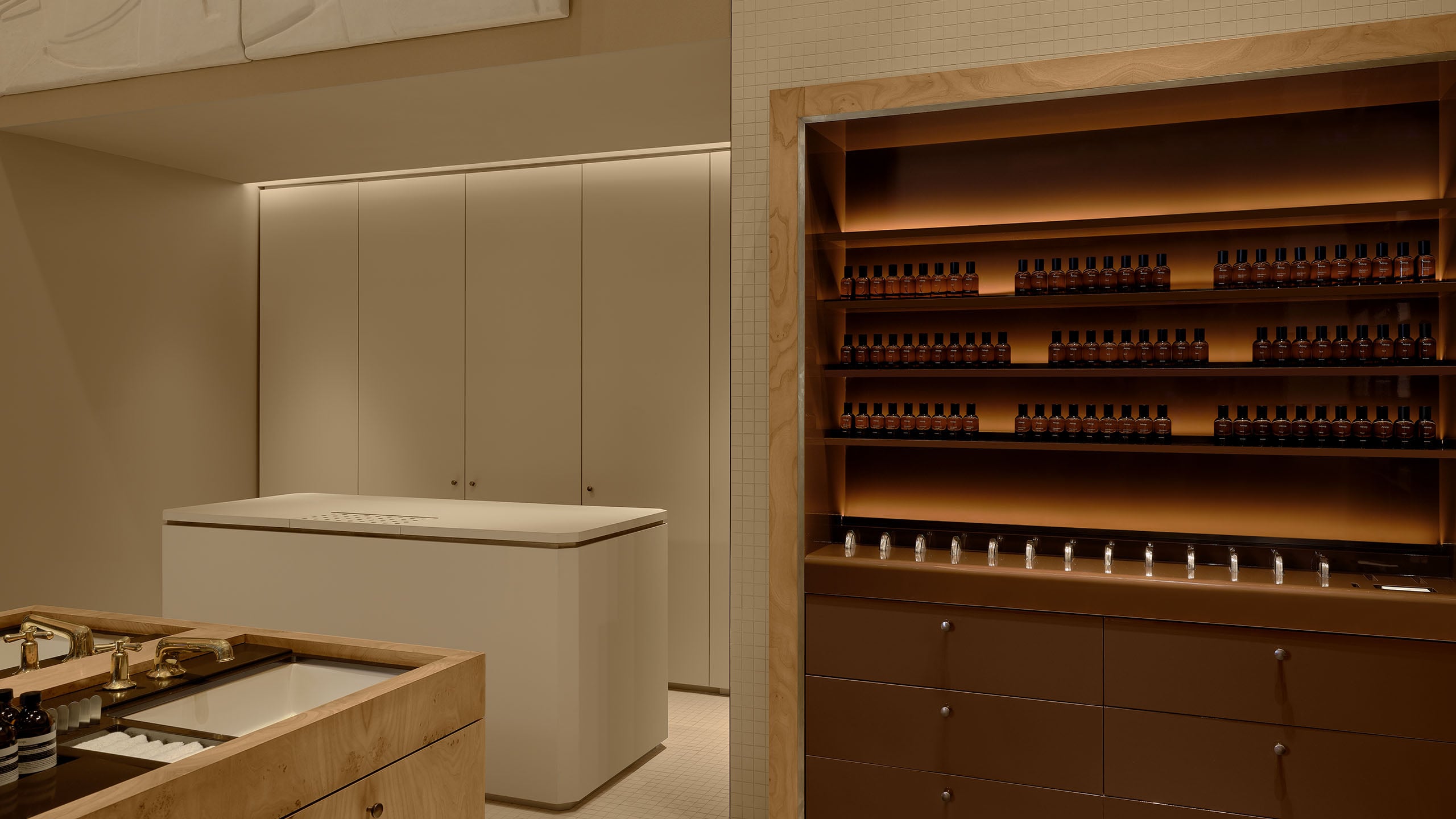 store interior featuring warmly lit brown product shelves, wooden center basin unit, and cream colored checkout counter against cream colored walls