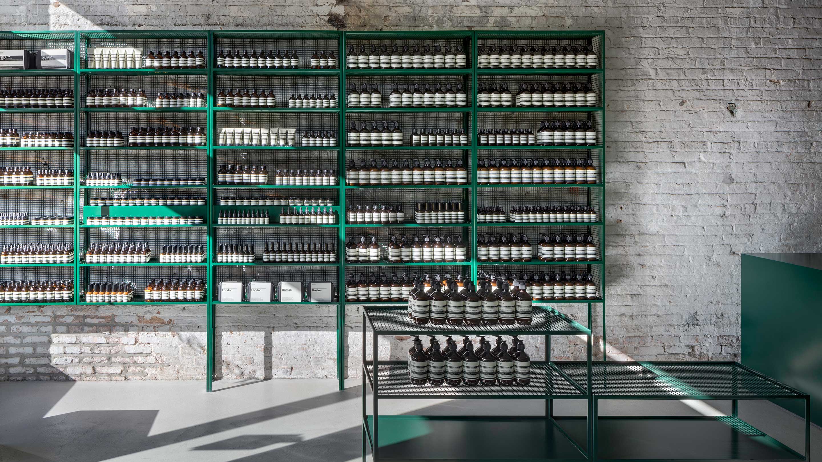 A light-filled store interior; a green, steel shelf holding Aesop product against a white brick wall. 