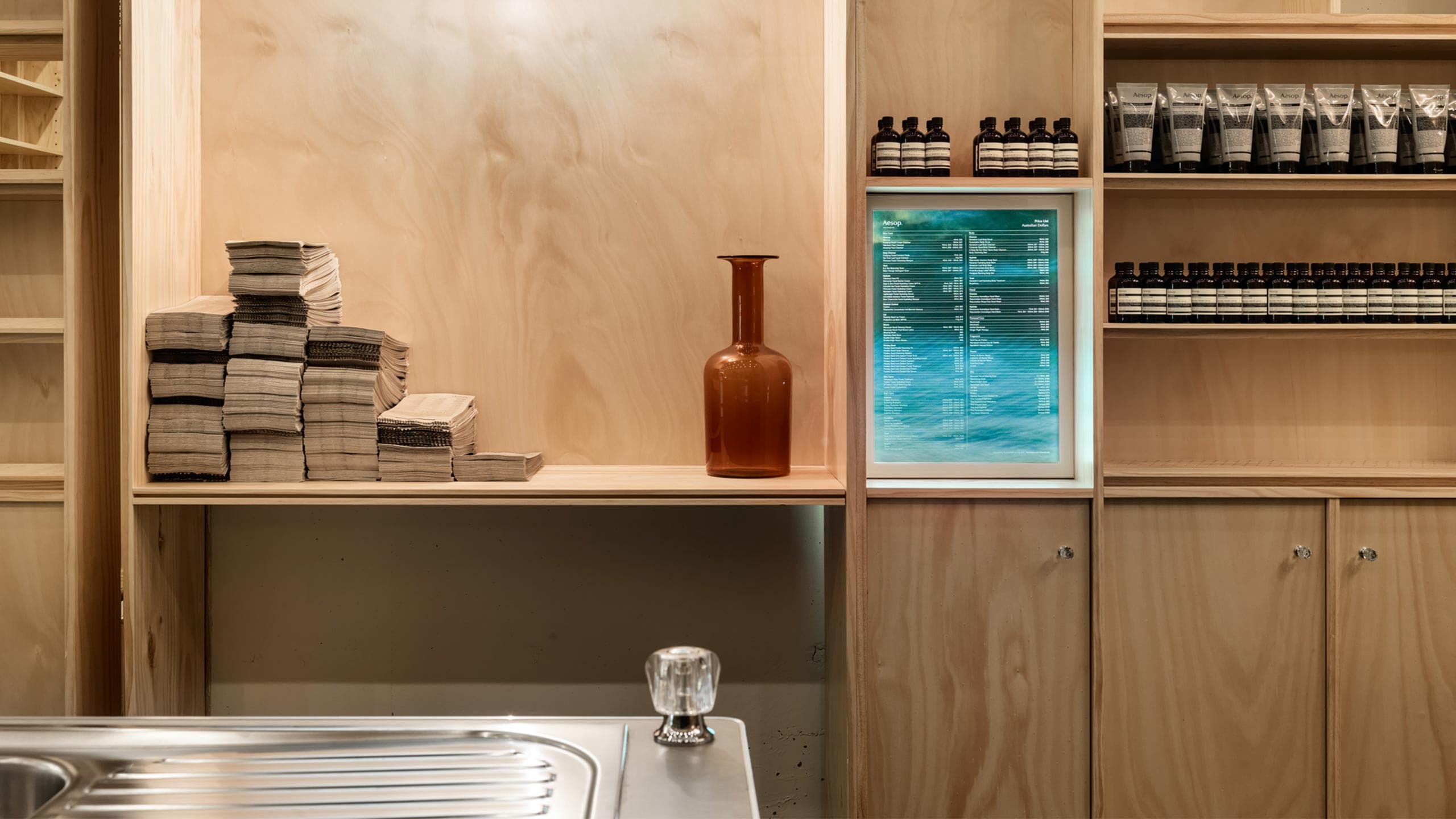 A stainless-steel sink, similar to those found in many Australian homes, is in the foreground. Timber shelving is in the background, adorned with products and an amber vase.
