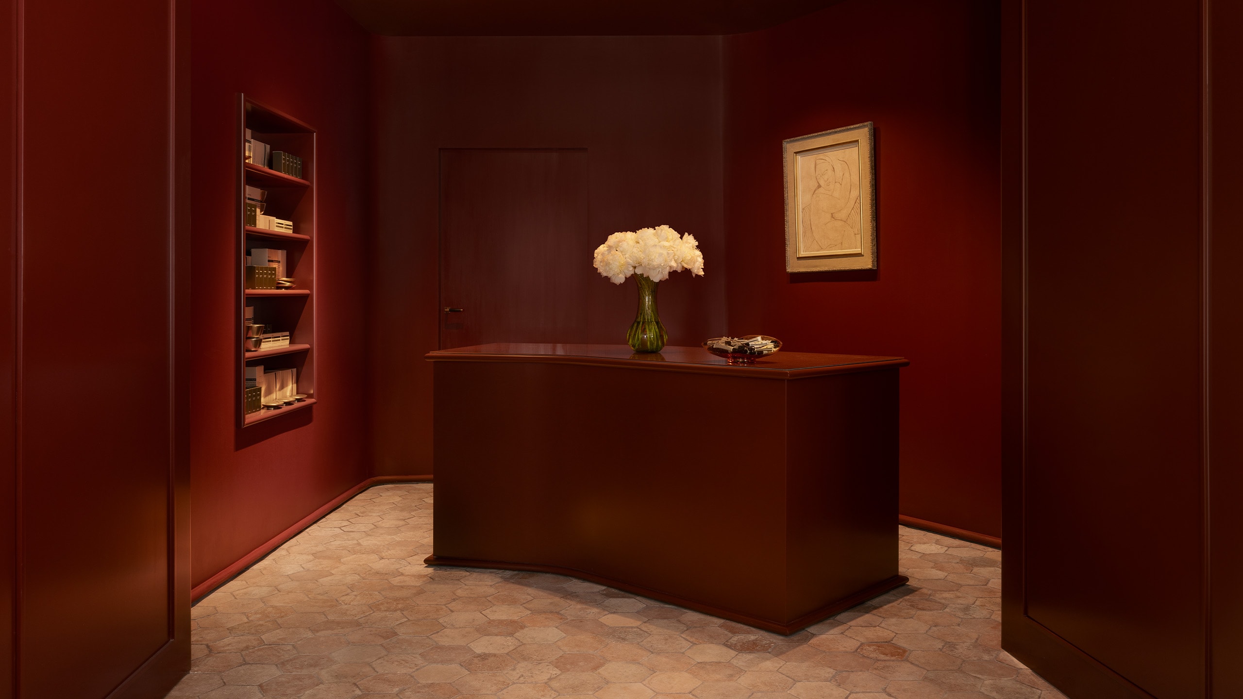 Aesop Avenue des Ternes store interior desk unit, painted a deep red, floral display in vase, mounted drawing and product display shelves.