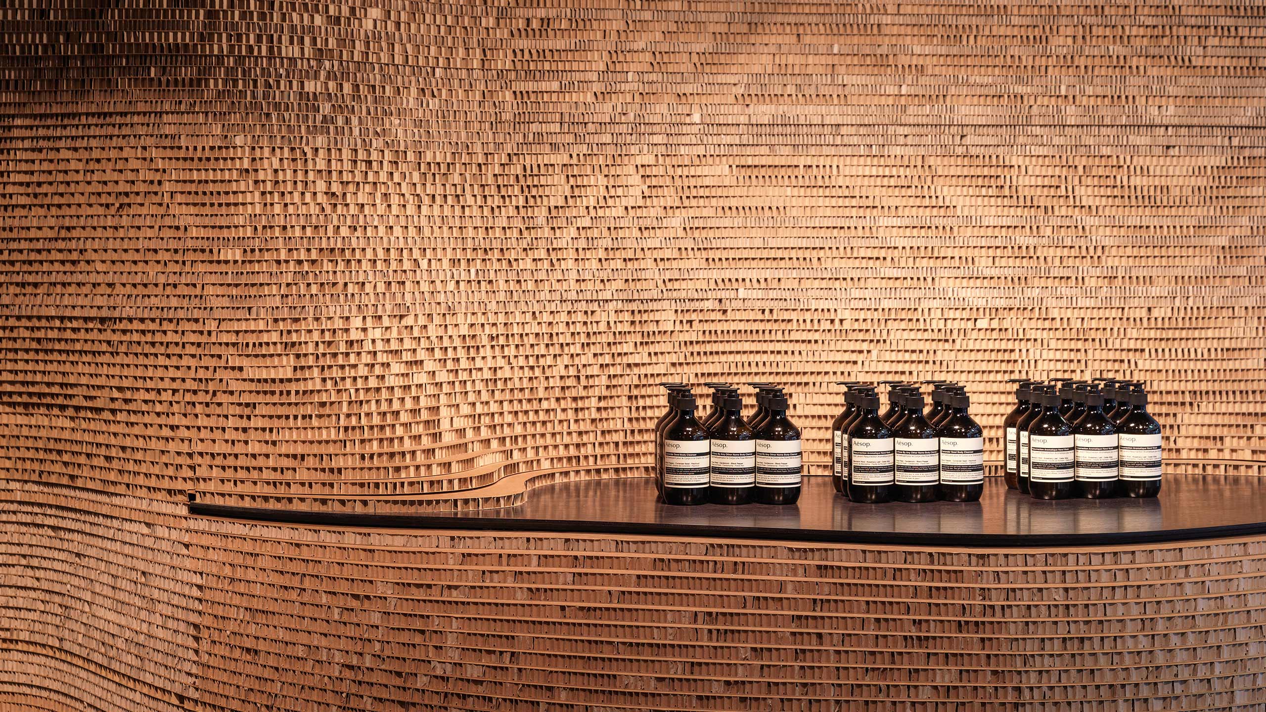 An oak shelf that is built into the curved cardboard wall. Twenty-seven black Aesop bottles rest on top of the curved shelf.