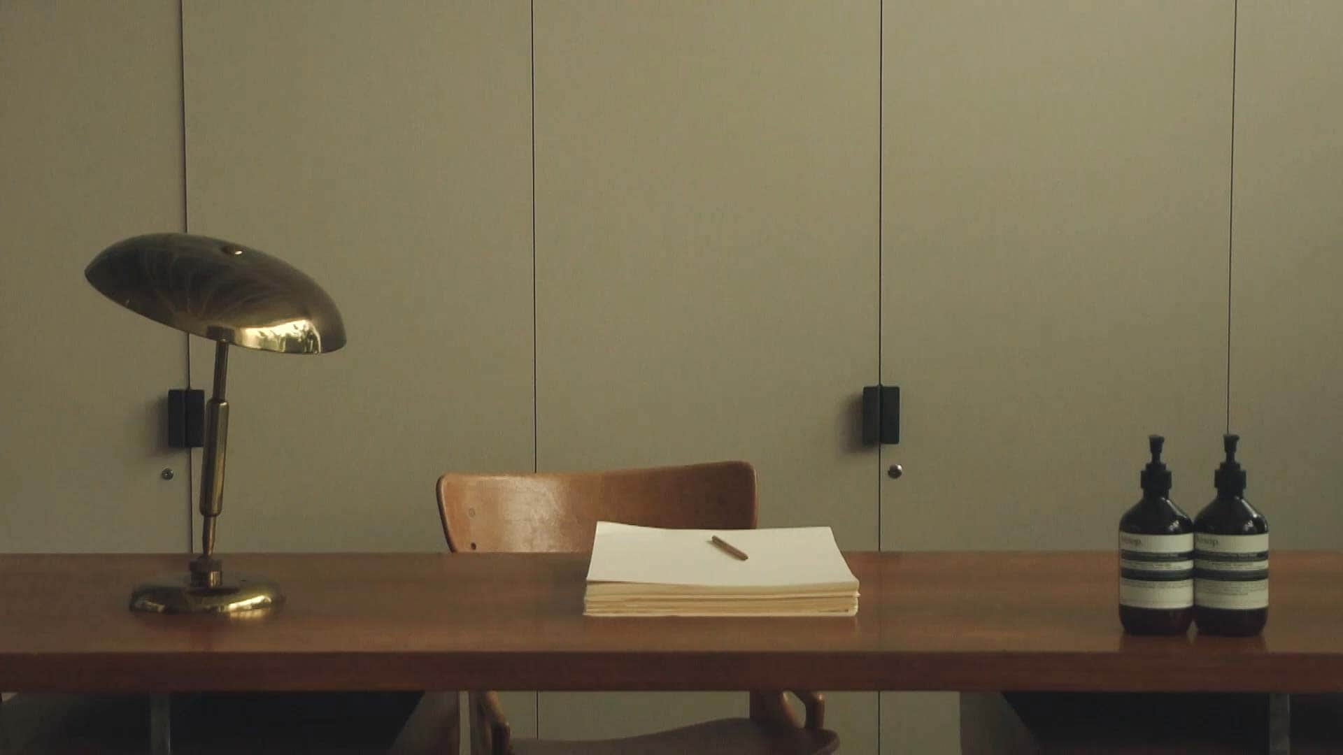 A pen and stack of paper on a wooden desk alongside a brass lamp and two bottles of Aesop hand balm.