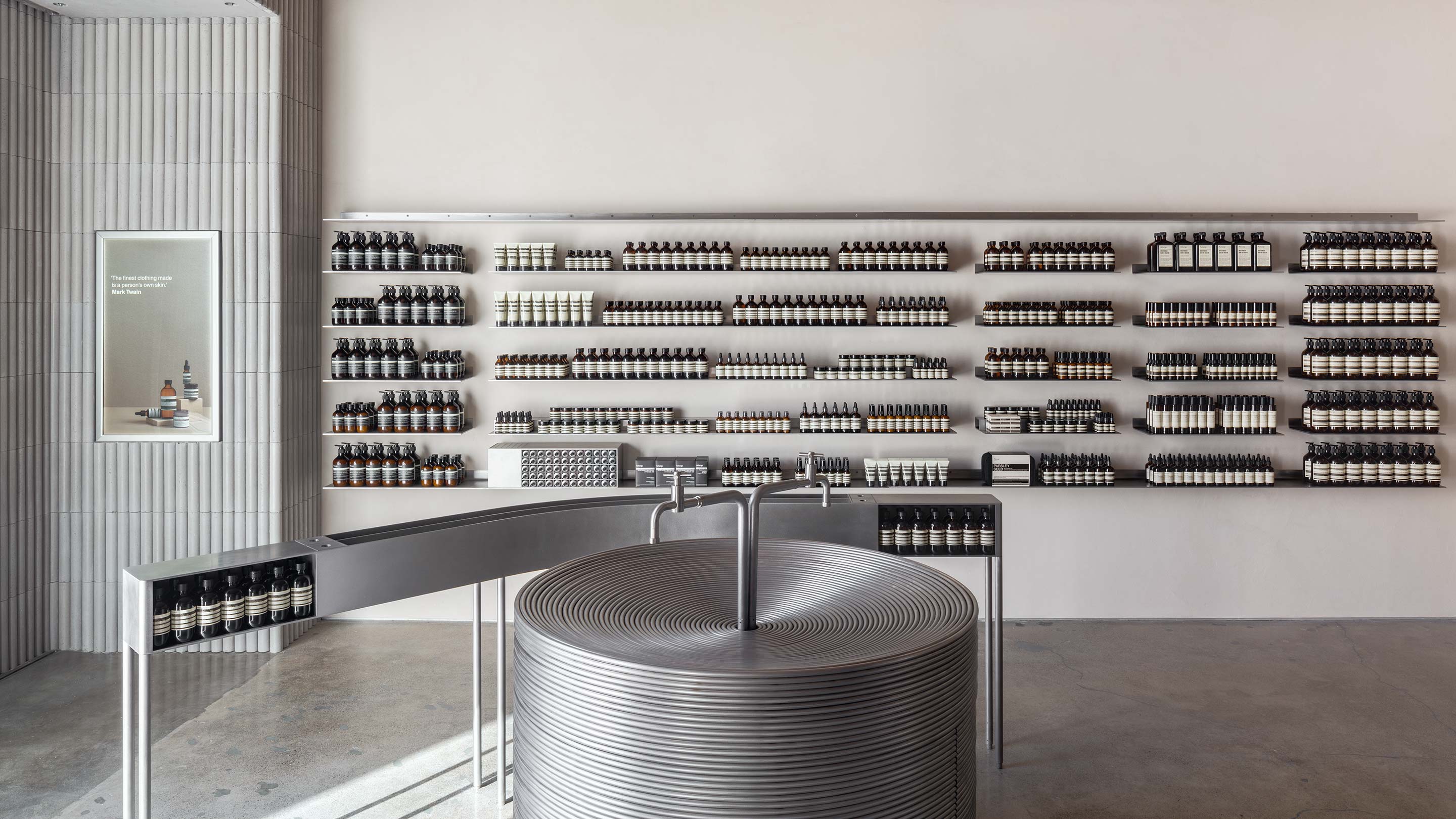 A circular sink made of brushed steel sits in the foreground; Aesop products arranged on shelves in the background.
