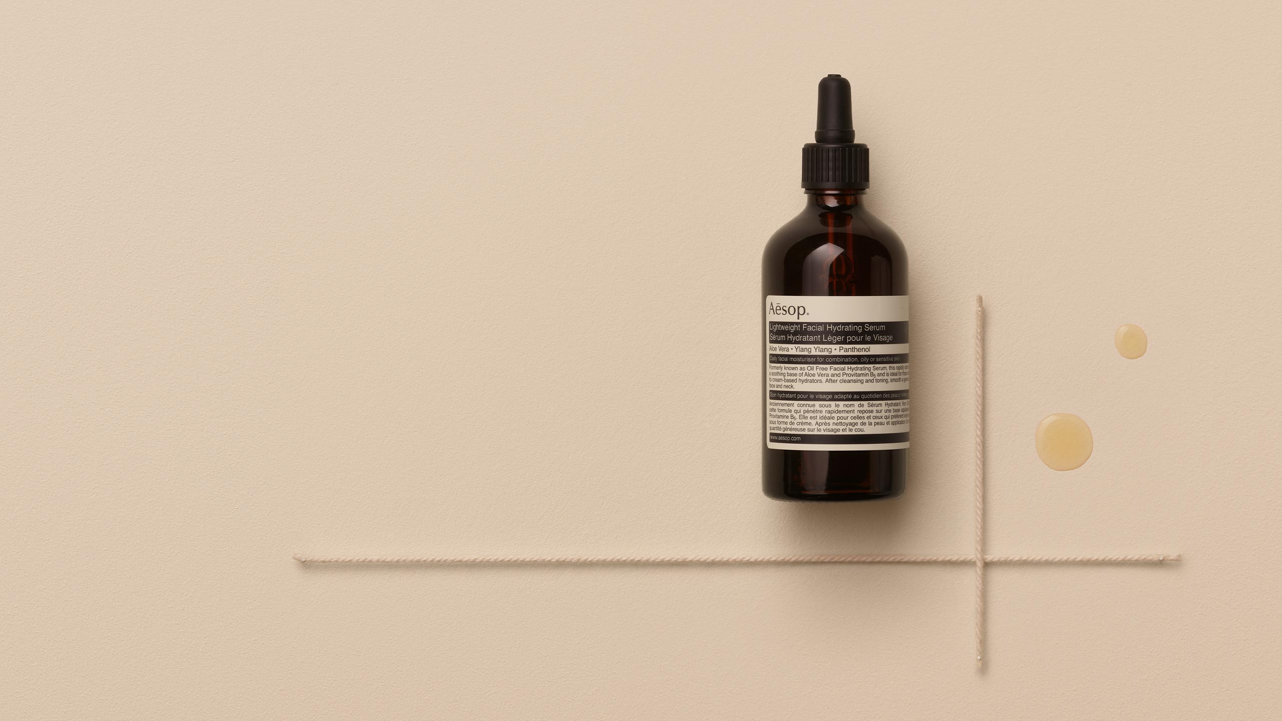 Aesop lightweight facial hydrating serum and two drops of the serum placed on textured beige background
